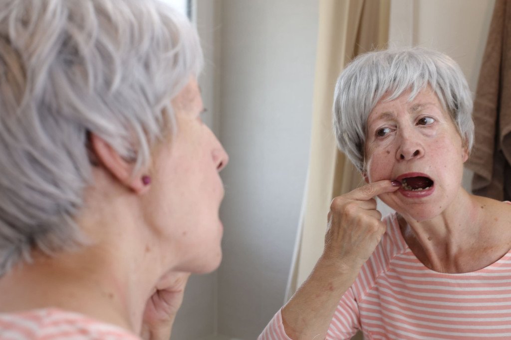 Une femme âgée inspectant sa dentition dans le miroir, possiblement en train de vérifier une prothèse dentaire ou un problème dentaire.