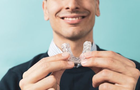 Un homme en costume noir et chemise rayée tenant et présentant une plaque occlusale transparente, un dispositif médical utilisé pour protéger les dents.