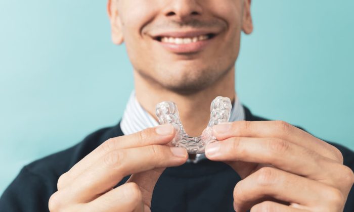 Un homme en costume noir et chemise rayée tenant et présentant une plaque occlusale transparente, un dispositif médical utilisé pour protéger les dents.